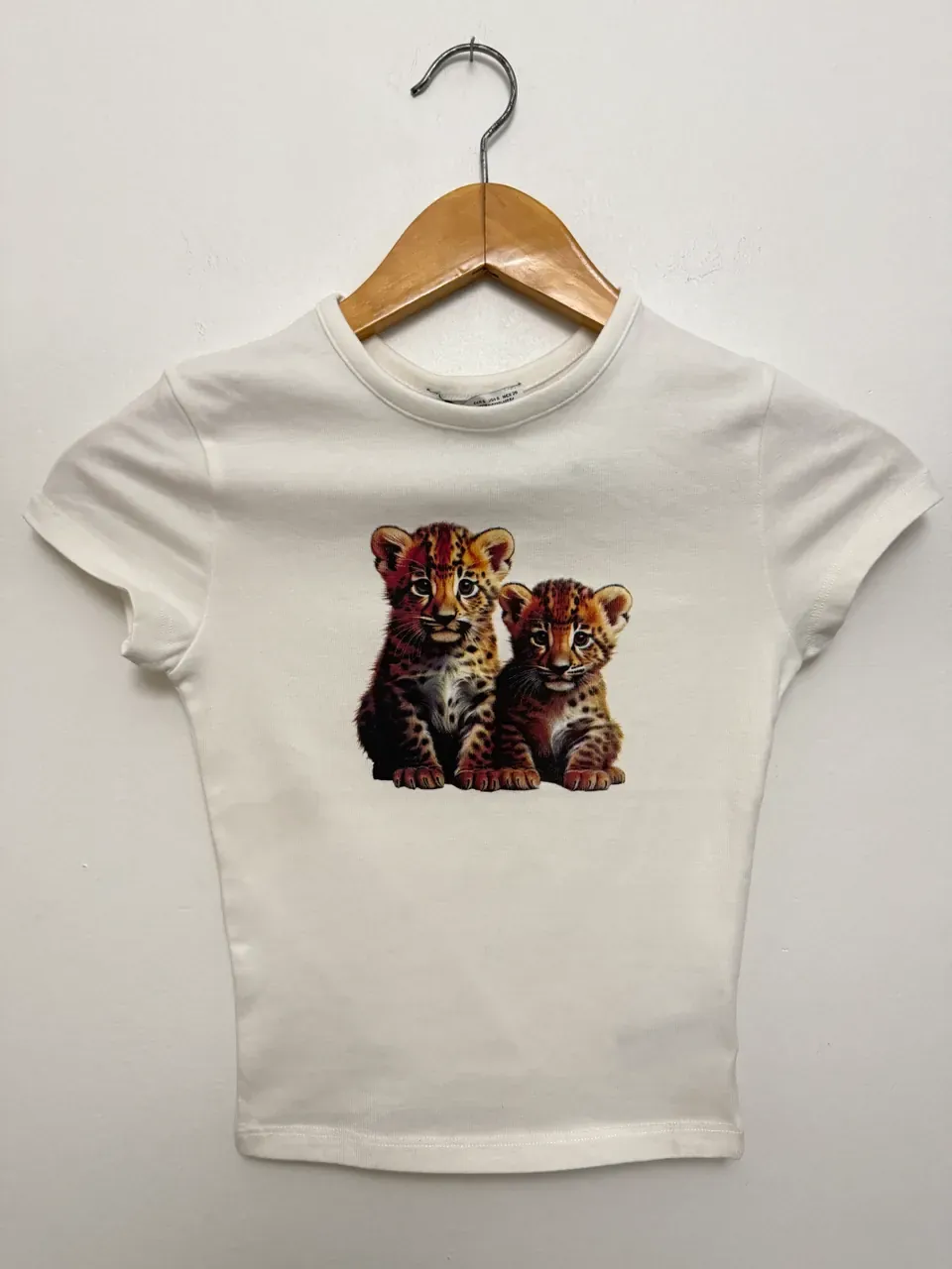 Baby tee blanca de manga corta con la estampa de dos cachorros de leopardo, colgada en una percha de madera