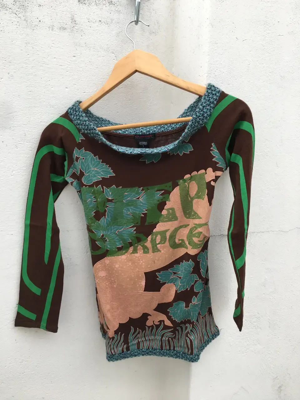 Remera de manga larga marrón con una estampa muy cargada de hojas y figuras en verde, celeste y rosa, y vivos verdes en las mangas, colgada en una percha